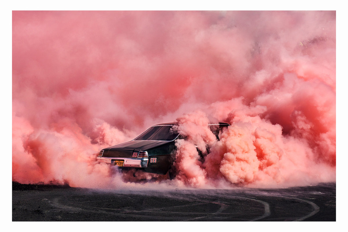 A black Holden chrome bumper sedan GTS bearing the number plate MCFRY performs a burnout, engulfed in a massive rolling cloud of vivid coral smoke that fills the entire frame from the burnout scared bitumen to the top of the image. The GTS badging and chrome rear bumper are just visible through the churning coral haze as smoke pours from both rear wheel arches across the rubber marked surface below. Fine art colour photographic print by Simon Davidson.
