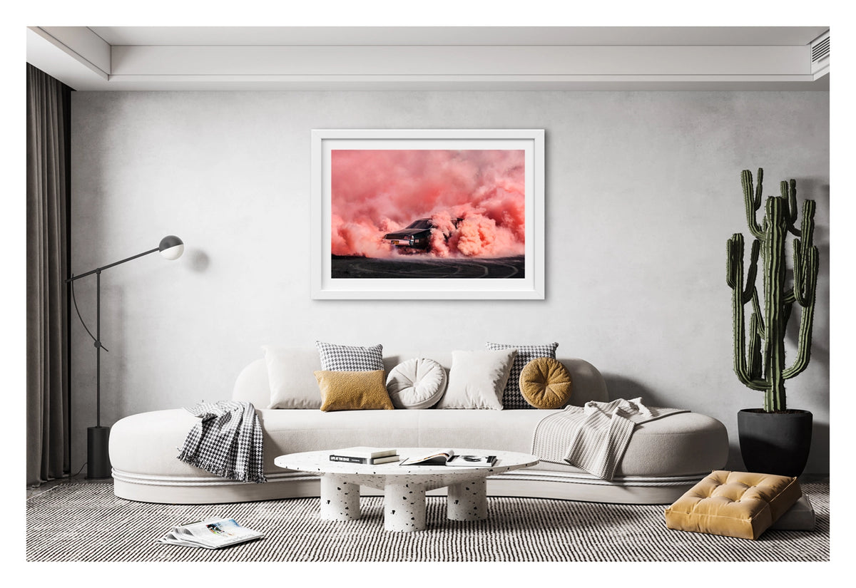 Framed in a white frame, shown in a contemporary living A black Holden chrome bumper sedan GTS bearing the number plate MCFRY performs a burnout, engulfed in a massive rolling cloud of vivid coral smoke that fills the entire frame from the burnout scared bitumen to the top of the image. The GTS badging and chrome rear bumper are just visible through the churning coral haze as smoke pours from both rear wheel arches across the rubber marked surface below. Fine art colour photographic print by Simon Davidson.