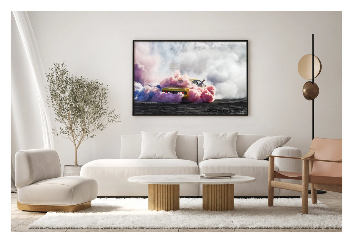 Framed in black, shown in a contemporary living space. A yellow Holden Commodore performs a burnout before a grandstand crowd, engulfed in a spectacular multicoloured cloud of pink, blue and white tyre smoke that swirls across the tyre marked black bitumen. A driver's arm reaches through the window with a single finger pointed skyward as the vibrant smoke rolls in every direction, the red tail lights glowing through the haze. Fine art colour photographic print by Simon Davidson.