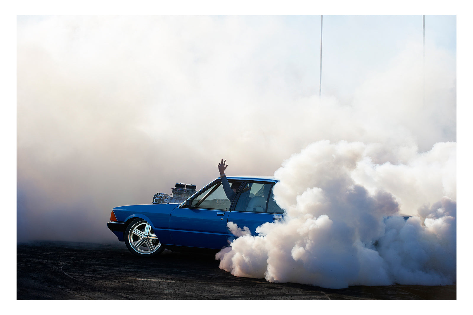 A blue Ford XD Falcon sedan with a large exposed blower engine mounted through the bonnet unleashes a burnout, a massive cloud of white tyre smoke engulfing the rear of the car and rolling across the tyre marked black bitumen. The passenger's arm reaches through the open window above the roofline as the bold blue body and polished five spoke wheels sit sharp against the billowing white smoke filling the frame. Fine art colour photographic print by Simon Davidson.