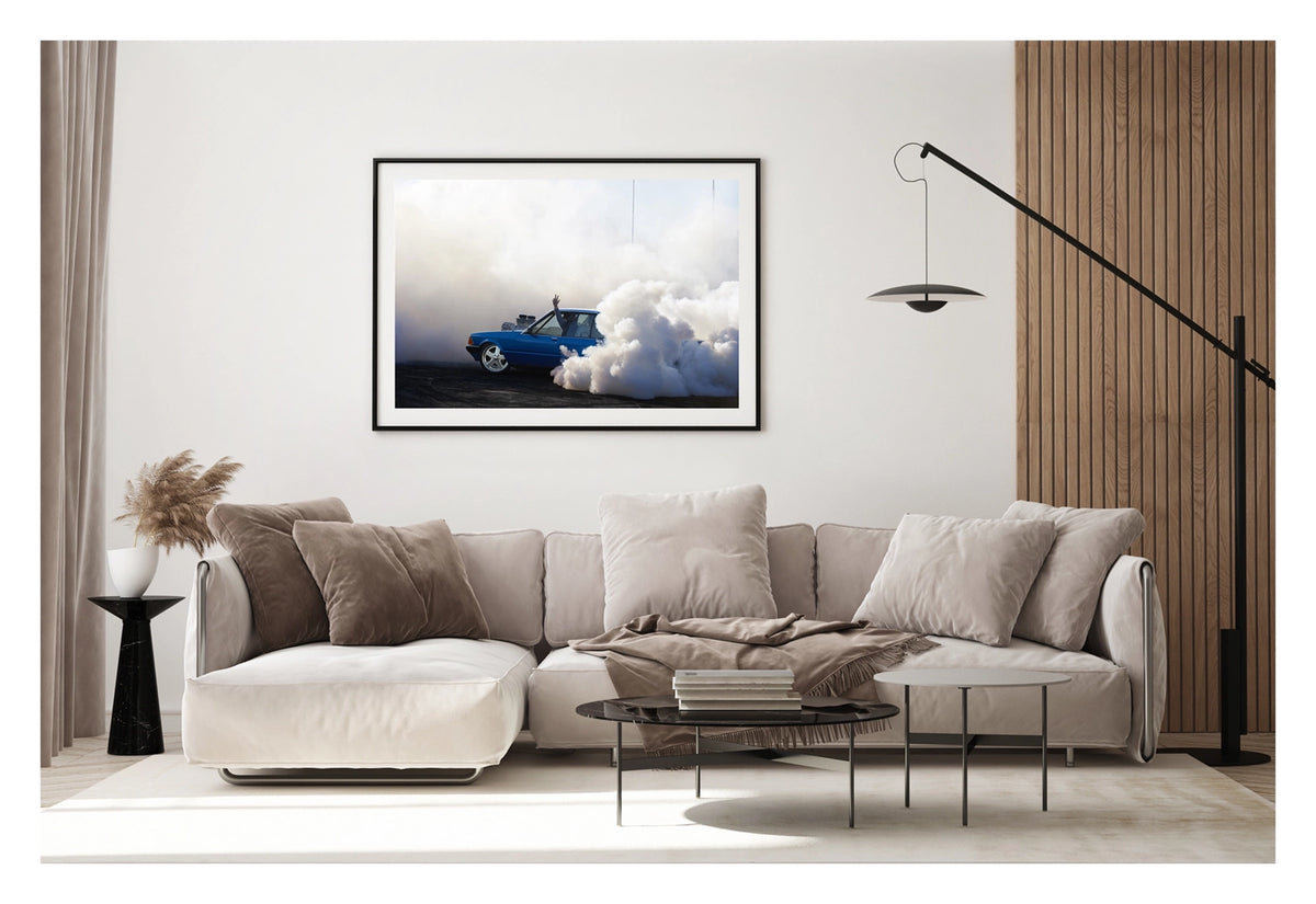 Framed in black, hanging in a contemporary space. A blue Ford XD Falcon sedan with a blown engine through the bonnet unleashes a burnout, a massive cloud of white tyre smoke engulfing the rear of the car and rolling across the tyre marked black bitumen. The passenger's arm reaches through the open window above the roofline as the bold blue body and polished five spoke wheels sit sharp against the billowing white smoke filling the frame. Fine art colour photographic print by Simon Davidson.