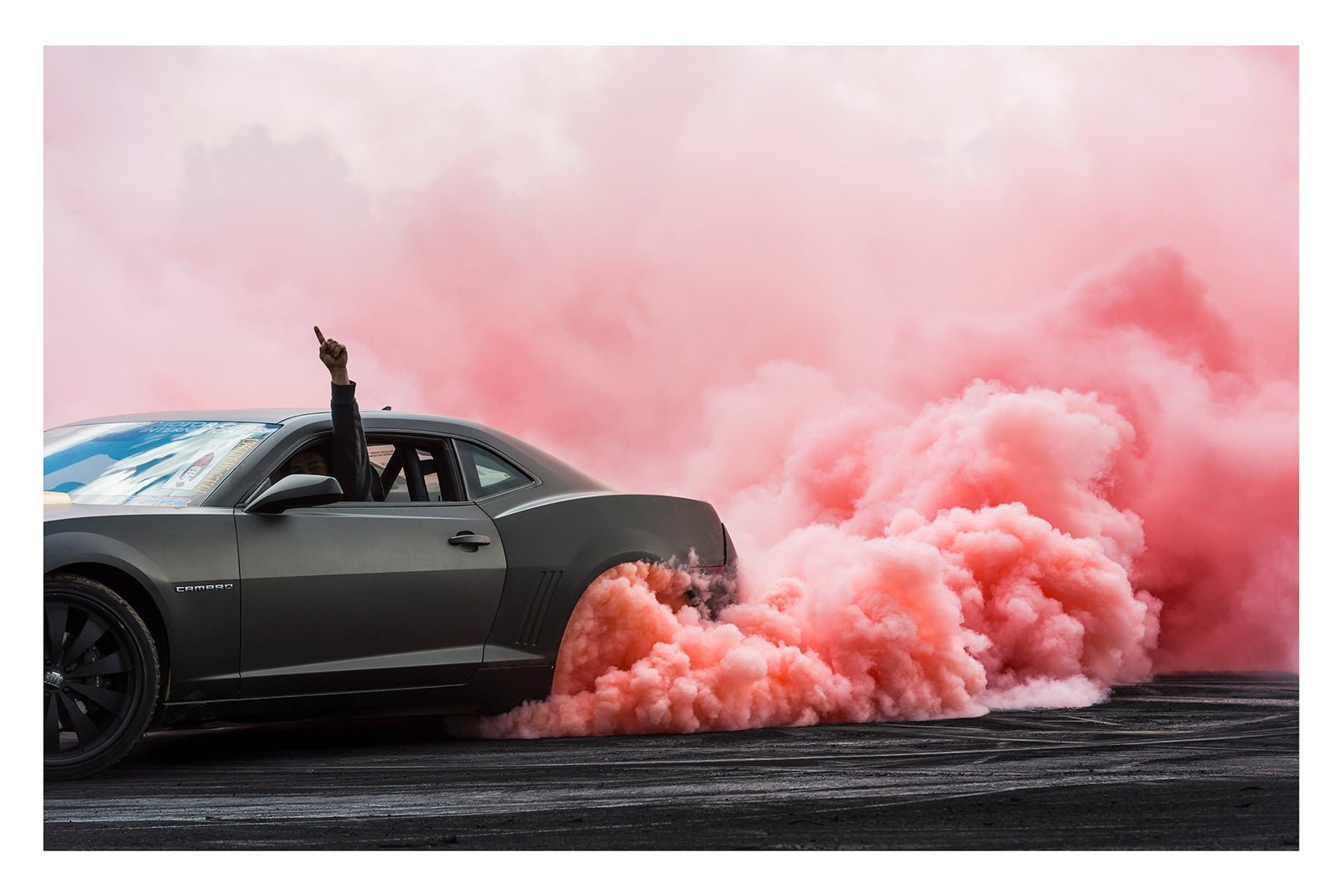 A modern matte black Chevrolet Camaro performs a burnout, a dense cloud of pink tyre smoke erupting from the rear wheel and billowing across the tyre damaged black bitumen. The passenger's arm reaches through the open window with a single finger pointed skyward as the bold contrast of the dark body against the rolling pink smoke fills the frame. Fine art colour photographic print by Simon Davidson.
