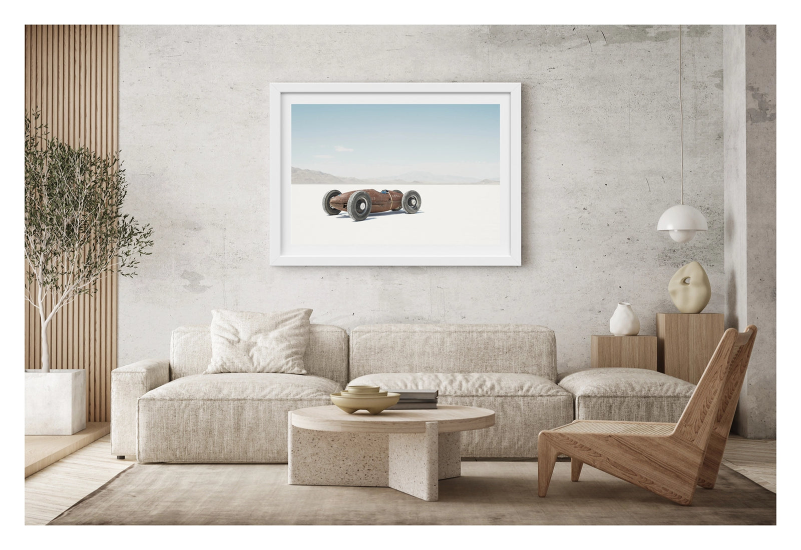 Framed in white. A small rust patinated vintage bellytanker landspeed race car sits alone on the blinding white salt of the Bonneville Salt Flats, its exposed wire wheels and torpedo shaped body giving it the appearance of a miniature prewar racing car. A hazy mountain range dissolves into the pale horizon behind it under a soft blue sky, the tiny machine dwarfed by the vast emptiness surrounding it. Fine art colour photographic print by Simon Davidson.