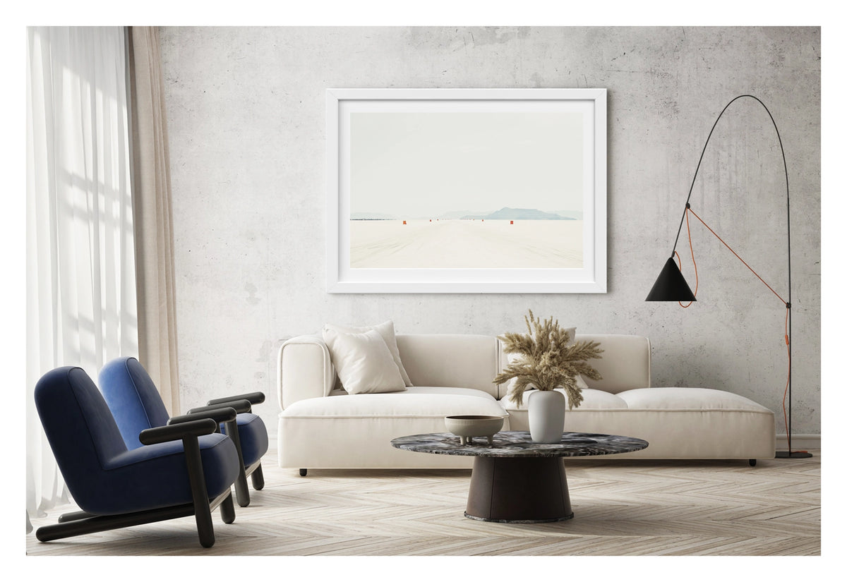 Framed in white, shown in a contemporary living space. We see a row of small orange course markers stretches into the vanishing point across the blinding white surface of the Bonneville Salt Flats, tyre tracks scoring the salt in the foreground as distant mountains dissolve into a pale grey horizon. The vast emptiness of the scene and the near seamless merging of salt flat and sky give the image a stark, minimalist beauty. Fine art colour photographic print by Simon Davidson.