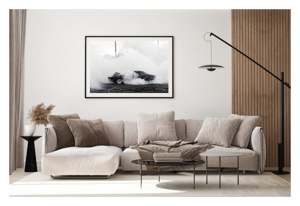 Framed in black, shown in a contemporary living space. We see a black Chevrolet Camaro with a polished supercharger protruding through the bonnet performs a burnout on a rubber streaked bitumen surface, engulfed in a thick cloud of white tyre smoke that erases the sky. Power lines and trees ghost through the haze in the background as the car sits at the centre of its own self made storm, wide slick tyres barely visible beneath the billowing smoke. Fine art colour photographic print by Simon Davidson.