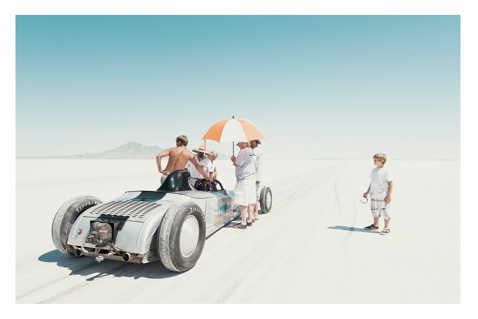 A vintage landspeed race car waits on the start line of the Bonneville salt flats as crew members shelter beneath an umbrella. A young boy looks on. The scene floats in the vast white silence of the Bonneville salt lake. Fine art photograph by Simon Davidson