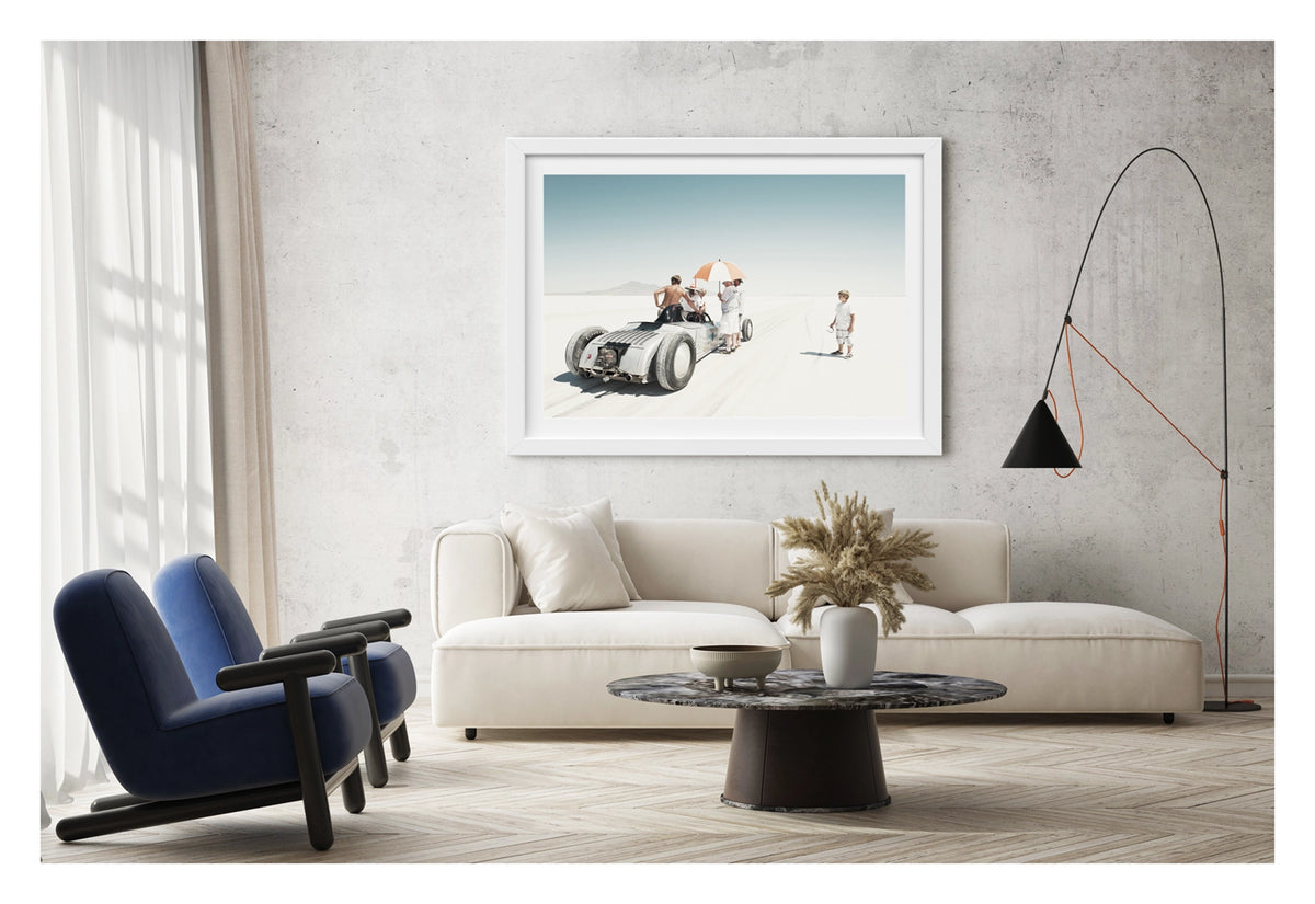 Framed in white, hanging in a contemporary space. We see a vintage landspeed race car waits on the start line of the Bonneville salt flats as crew members shelter beneath an umbrella. A young boy looks on. The scene floats in the vast white silence of the Bonneville salt lake. Fine art photograph by Simon Davidson