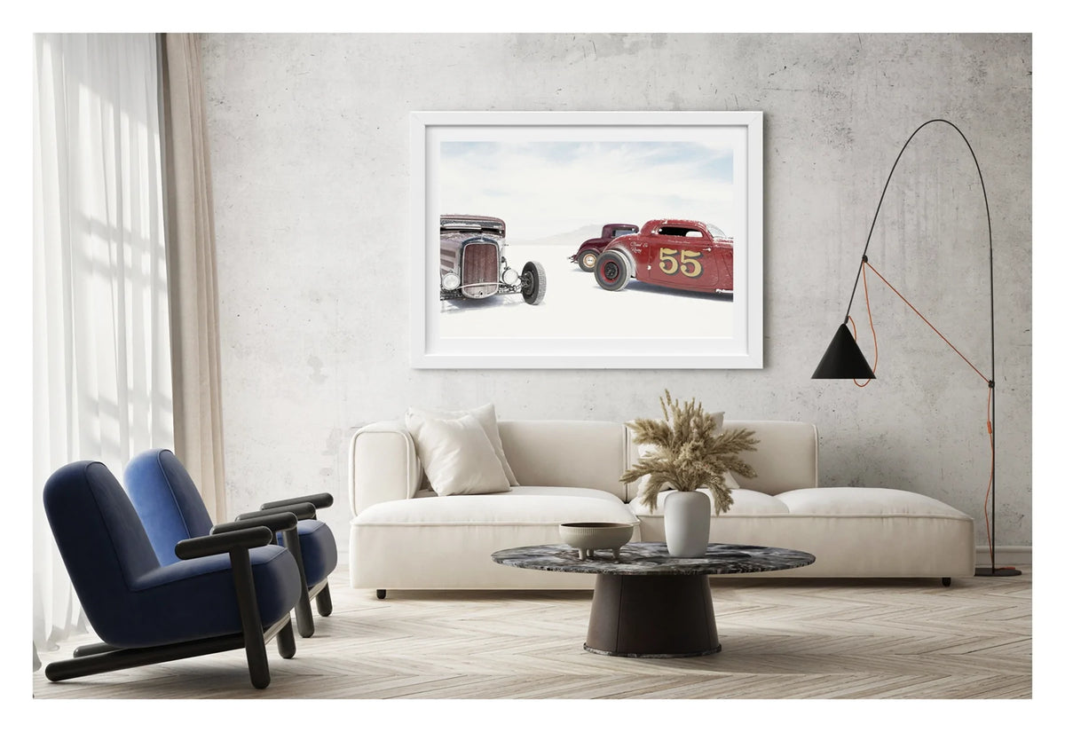 Framed in a white on a concrete wall. Two salt encrusted Rolling Bones hot rods sit on the blinding white surface of the Bonneville Salt Flats, their weathered bodies dusted with salt crystal residue. A third maroon coupe visible between them in the middle distance. A pale mountain range dissolves into the white horizon behind them. Fine art colour photographic print by Simon Davidson.