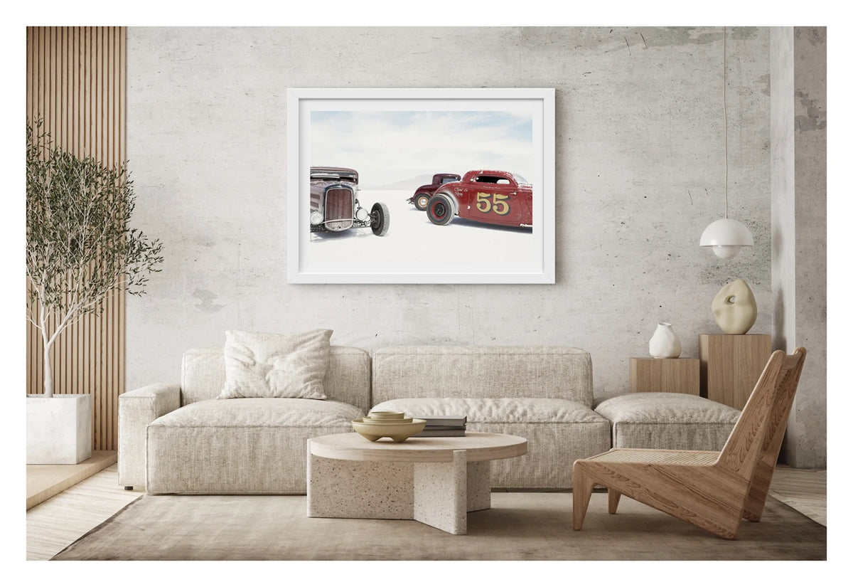 Framed in a white on a concrete wall. Two salt encrusted Rolling Bones hot rods sit on the blinding white surface of the Bonneville Salt Flats, their weathered bodies dusted with salt crystal residue. A third maroon coupe visible between them in the middle distance. A pale mountain range dissolves into the white horizon behind them. Fine art colour photographic print by Simon Davidson.