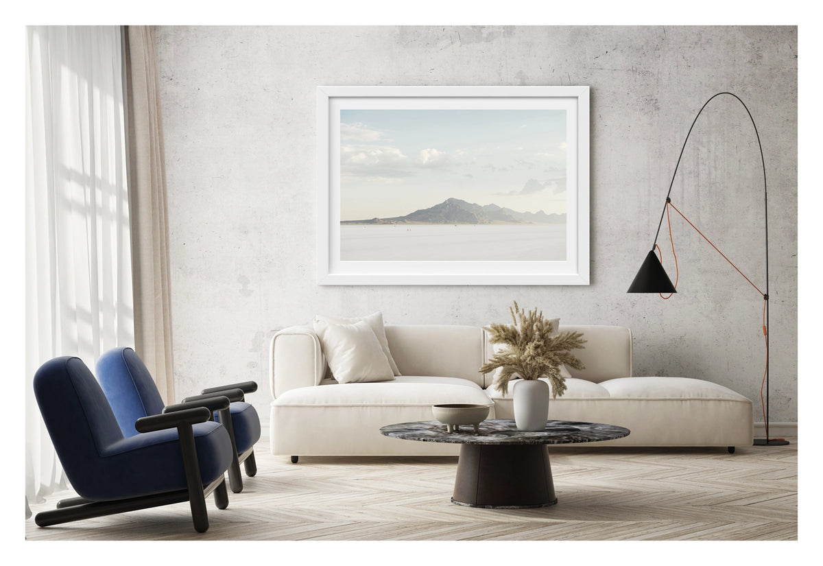 Framed in white, hanging in a contemporary living space. An image of the vast expanse of the Bonneville Salt Flats stretches towards the Silver Mountains, it's softened by haze beneath a pale blue sky of billowing cumulus clouds. Tiny orange cones dot the salt at the base of the mountain, their miniature scale emphasising the immense stillness and silence of the landscape. Fine art colour photographic print by Simon Davidson.