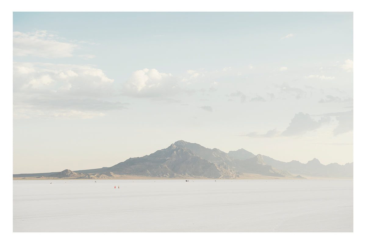 An image of the vast expanse of the Bonneville Salt Flats stretches towards the Silver Mountains, it's softened by haze beneath a pale blue sky of billowing cumulus clouds. Tiny orange cones dot the salt at the base of the mountain, their miniature scale emphasising the immense stillness and silence of the landscape. Fine art colour photographic print by Simon Davidson.