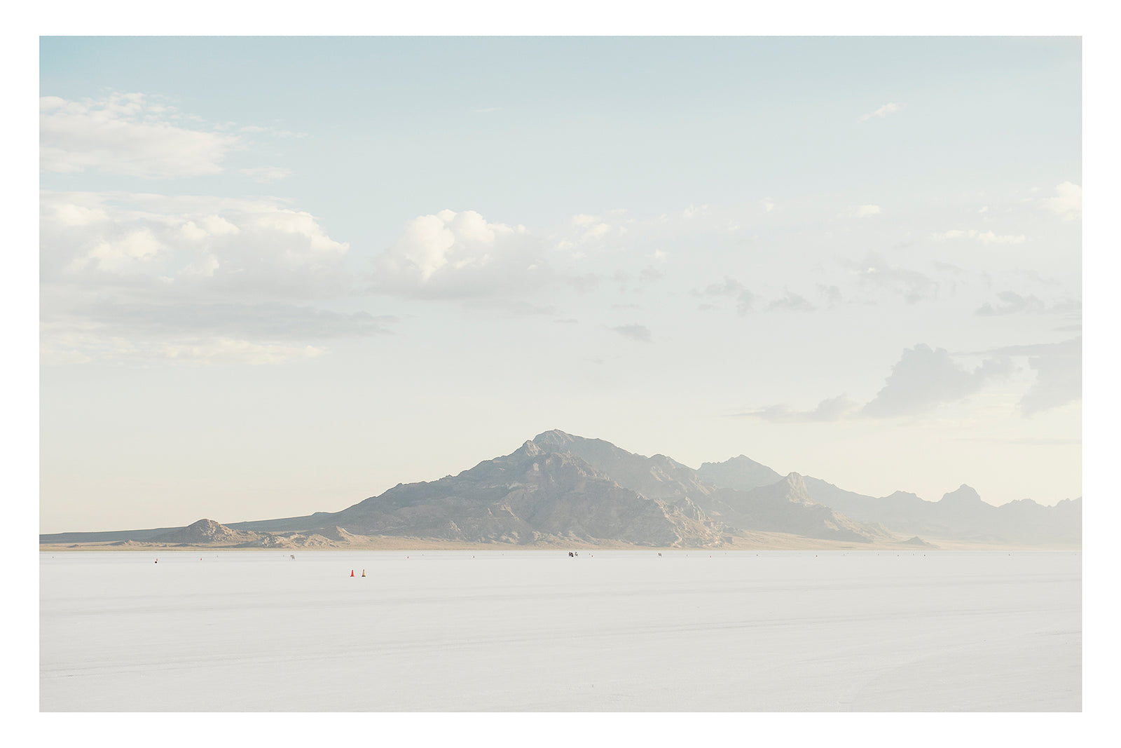 An image of the vast expanse of the Bonneville Salt Flats stretches towards the Silver Mountains, it's softened by haze beneath a pale blue sky of billowing cumulus clouds. Tiny orange cones dot the salt at the base of the mountain, their miniature scale emphasising the immense stillness and silence of the landscape. Fine art colour photographic print by Simon Davidson.