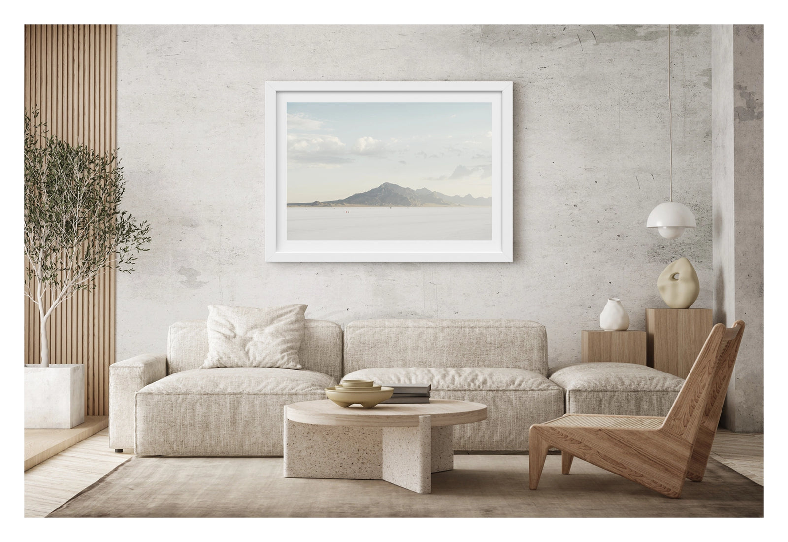 Framed in white, hanging in a contemporary living space. An image of the vast expanse of the Bonneville Salt Flats stretches towards the Silver Mountains, it's softened by haze beneath a pale blue sky of billowing cumulus clouds. Tiny orange cones dot the salt at the base of the mountain, their miniature scale emphasising the immense stillness and silence of the landscape. Fine art colour photographic print by Simon Davidson.