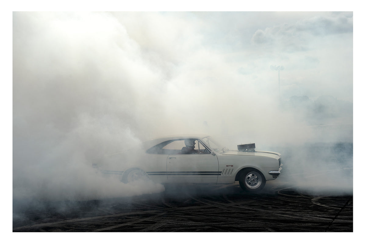 A cream Holden HK Monaro GTS with the engine protruding through the bonnet plants a burnout on heavily rubber marked black bitumen, the car emerging from a vast sea of white tyre smoke that fills the frame from ground to sky. The classic fastback roofline, GTS side stripes and the helmeted driver are just visible through the swirling haze as smoke rolls in every direction across the dark surface below. Fine art colour photographic print by Simon Davidson.