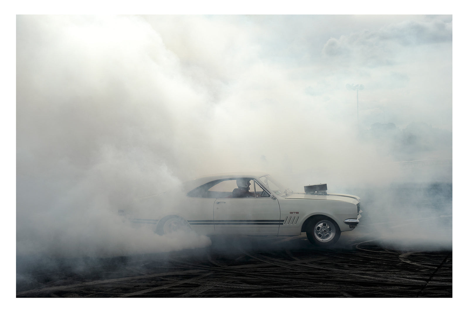 A cream Holden HK Monaro GTS with the engine protruding through the bonnet plants a burnout on heavily rubber marked black bitumen, the car emerging from a vast sea of white tyre smoke that fills the frame from ground to sky. The classic fastback roofline, GTS side stripes and the helmeted driver are just visible through the swirling haze as smoke rolls in every direction across the dark surface below. Fine art colour photographic print by Simon Davidson.