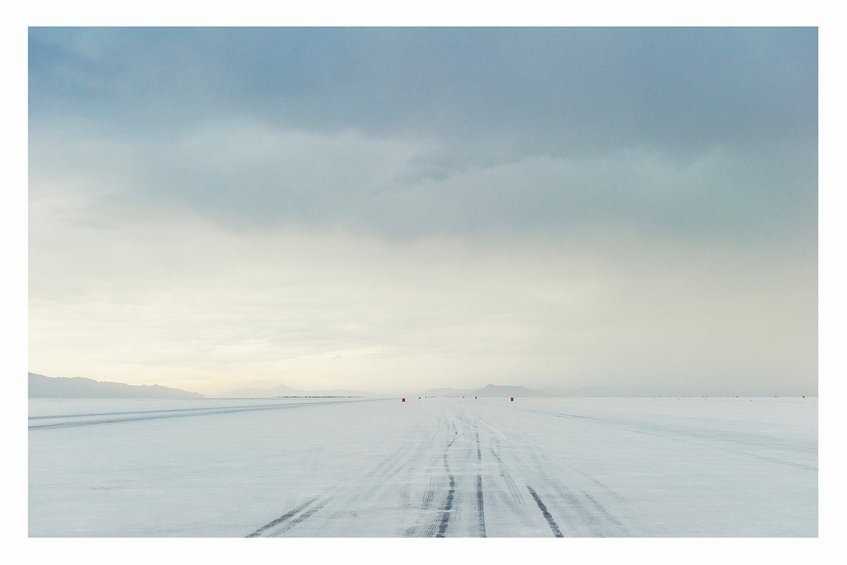 The No.1 race track of the Bonneville Salt Flats stretches to the vanishing point under a heavy brooding storm sky, tyre tracks scoring the salt surface as a row of small red timing markers disappear into the horizon toward a distant mountain range that's glowing with pale light. The threatening grey sky and luminous horizon create a dramatic sense of foreboding over the otherwise empty and silent expanse. Fine art colour photographic print by Simon Davidson.