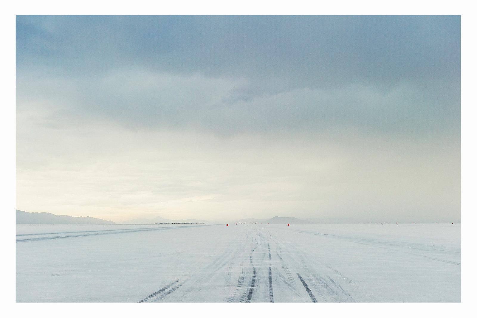 The No.1 race track of the Bonneville Salt Flats stretches to the vanishing point under a heavy brooding storm sky, tyre tracks scoring the salt surface as a row of small red timing markers disappear into the horizon toward a distant mountain range that's glowing with pale light. The threatening grey sky and luminous horizon create a dramatic sense of foreboding over the otherwise empty and silent expanse. Fine art colour photographic print by Simon Davidson.