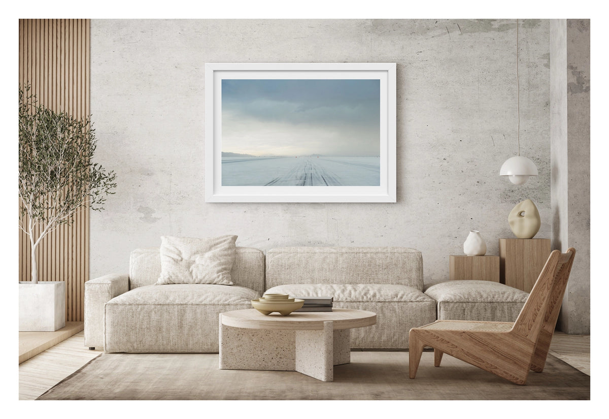 Framed in white, hanging in a contemporary living space. We see the No.1 race track of the Bonneville Salt Flats stretches to the vanishing point under a heavy brooding storm sky, tyre tracks scoring the salt surface as a row of small red timing markers disappear into the horizon toward a distant mountain range. The threatening grey sky and luminous horizon create a dramatic sense of foreboding over the otherwise empty and silent expanse. Fine art colour photographic print by Simon Davidson.