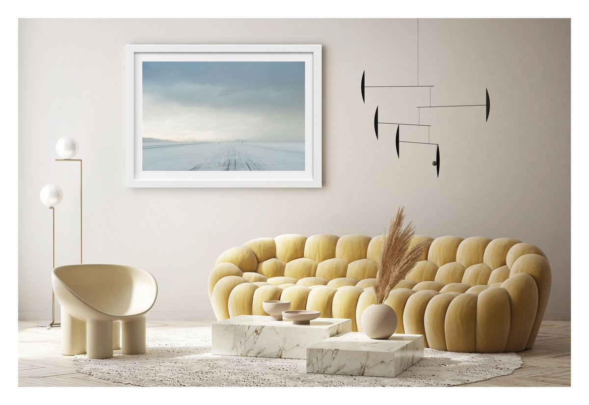 Framed in white, hanging in a contemporary living space. We see the No.1 race track of the Bonneville Salt Flats stretches to the vanishing point under a heavy brooding storm sky, tyre tracks scoring the salt surface as a row of small red timing markers disappear into the horizon toward a distant mountain range. The threatening grey sky and luminous horizon create a dramatic sense of foreboding over the otherwise empty and silent expanse. Fine art colour photographic print by Simon Davidson.