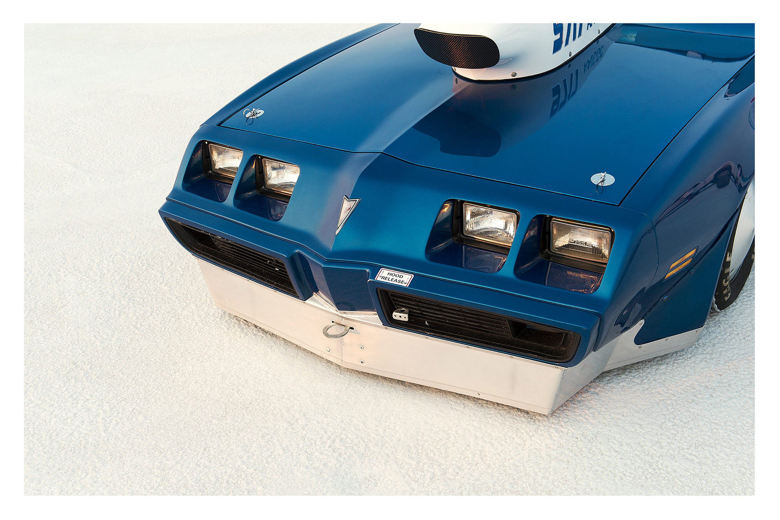 A close up of the front end of a blue Pontiac Trans Am land speed racer sitting low on the white salt of the Bonneville Salt Flats, its distinctive quad headlights, Pontiac arrow nose and fabricated aluminium lower valance filling the frame. A streamliner style intake is visible above the bonnet line, the deep blue paint and salt crusted surface contrasting sharply in the crisp flat light. Fine art colour photographic print by Simon Davidson.