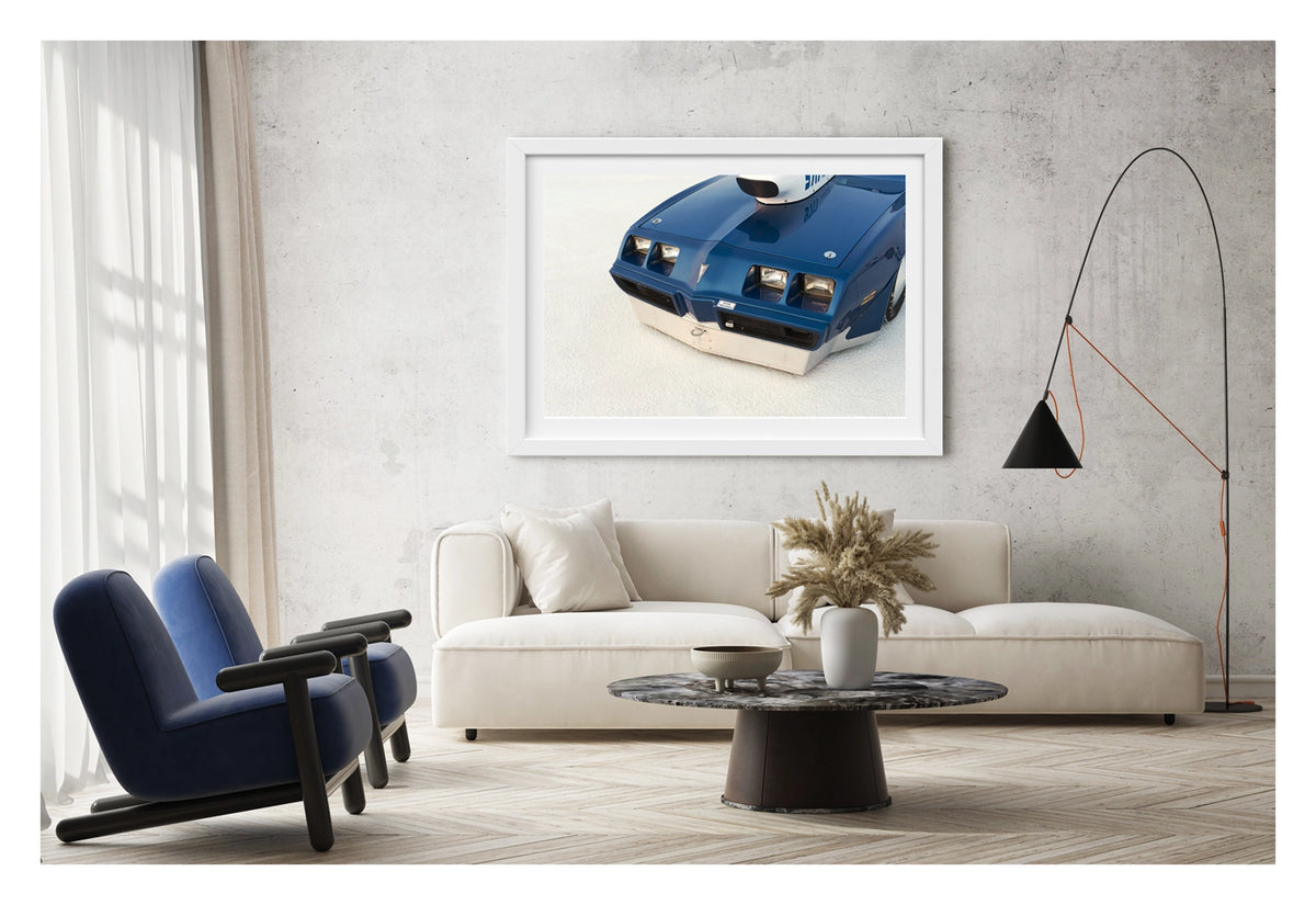 Framed in white, hanging in a contemporary living space. We see a close up of the front end of a blue Pontiac Trans Am land speed racer sitting low on the white salt of the Bonneville Salt Flats, its distinctive quad headlights, Pontiac arrow nose and fabricated aluminium lower valance filling the frame. A streamliner style intake is visible above the bonnet line, the deep blue paint and salt crusted surface contrasting sharply in the crisp flat light. Fine art colour photographic print by Simon Davidson.