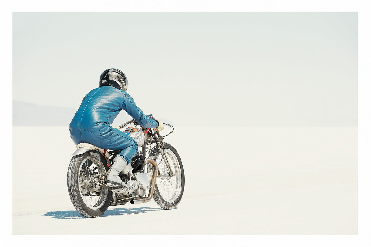 A lone motorcycle racer dressed in a blue leather suit and silver helmet rides a vintage Triumph, land speed racing motorcycle across the blinding white expanse of the Bonneville salt flats, the rider and bike reduced to a small solitary figure against an almost entirely white horizon and washed out sky. Fine art colour photographic print by Simon Davidson.