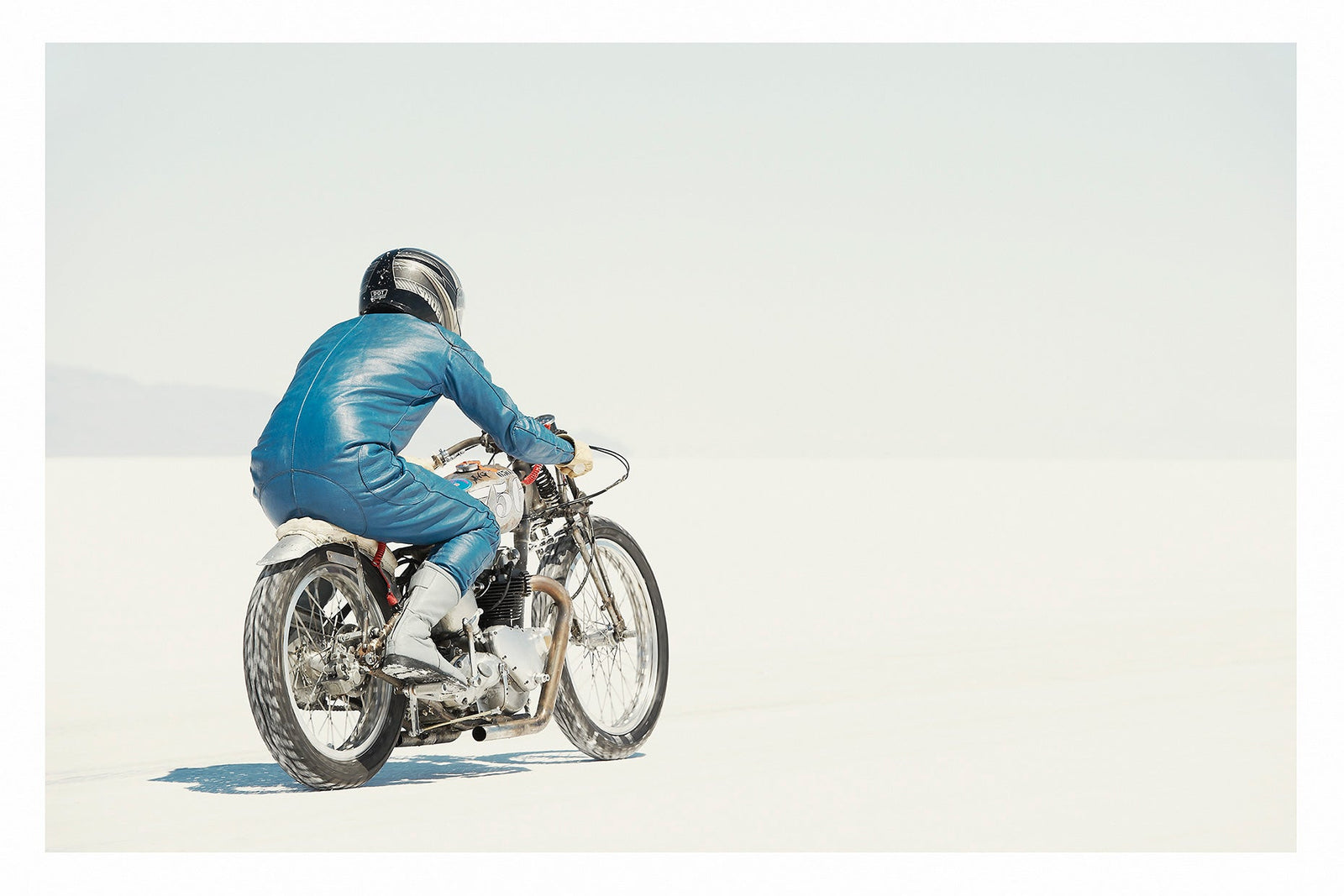 A lone motorcycle racer dressed in a blue leather suit and silver helmet rides a vintage Triumph, land speed racing motorcycle across the blinding white expanse of the Bonneville salt flats, the rider and bike reduced to a small solitary figure against an almost entirely white horizon and washed out sky. Fine art colour photographic print by Simon Davidson.