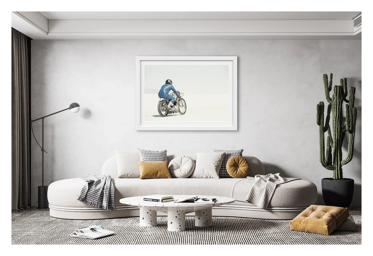 Framed in white. A lone motorcycle racer dressed in a blue leather suit and silver helmet rides a vintage Triumph, land speed racing motorcycle across the blinding white expanse of the Bonneville salt flats, the rider and bike reduced to a small solitary figure against an almost entirely white horizon and washed out sky. Shown in a contemporary living room. Fine art colour photographic print by Simon Davidson.