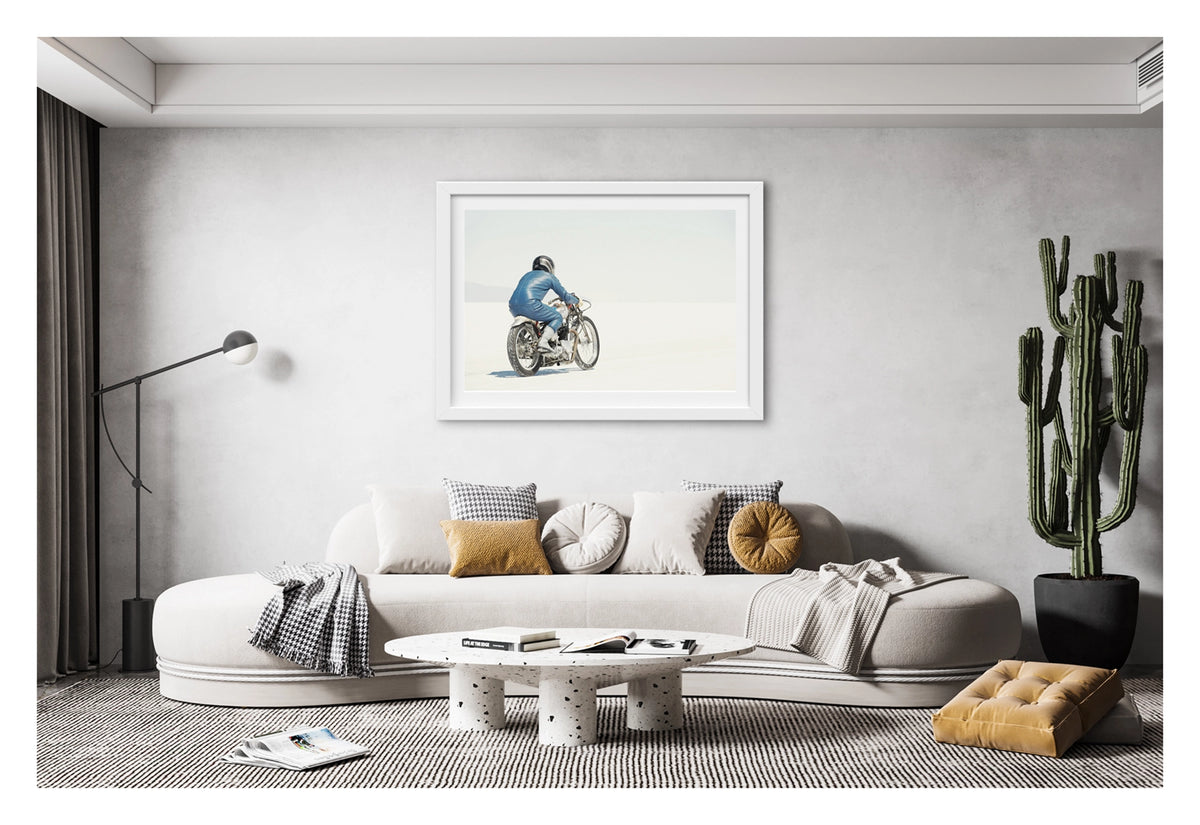 Framed in white, hanging in a contemporary living space. An image of a  lone motorcycle racer dressed in a blue leather suit and silver helmet rides a vintage Triumph, land speed racing motorcycle across the blinding white expanse of the Bonneville salt flats, the rider and bike reduced to a small solitary figure against an almost entirely white horizon and washed out sky. Shown in a contemporary living room. Fine art colour photographic print by Simon Davidson.