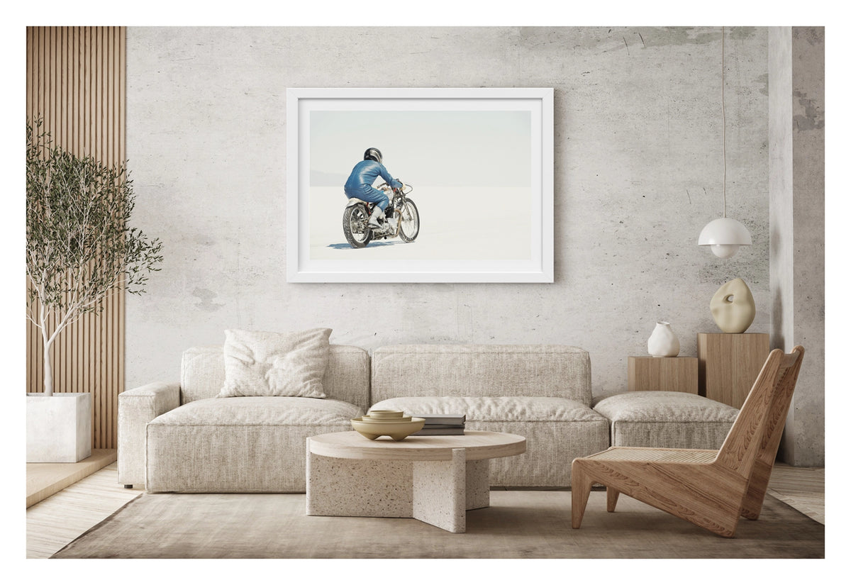 Framed in white, hanging in a contemporary living space. An image of a  lone motorcycle racer dressed in a blue leather suit and silver helmet rides a vintage Triumph, land speed racing motorcycle across the blinding white expanse of the Bonneville salt flats, the rider and bike reduced to a small solitary figure against an almost entirely white horizon and washed out sky. Shown in a contemporary living room. Fine art colour photographic print by Simon Davidson.