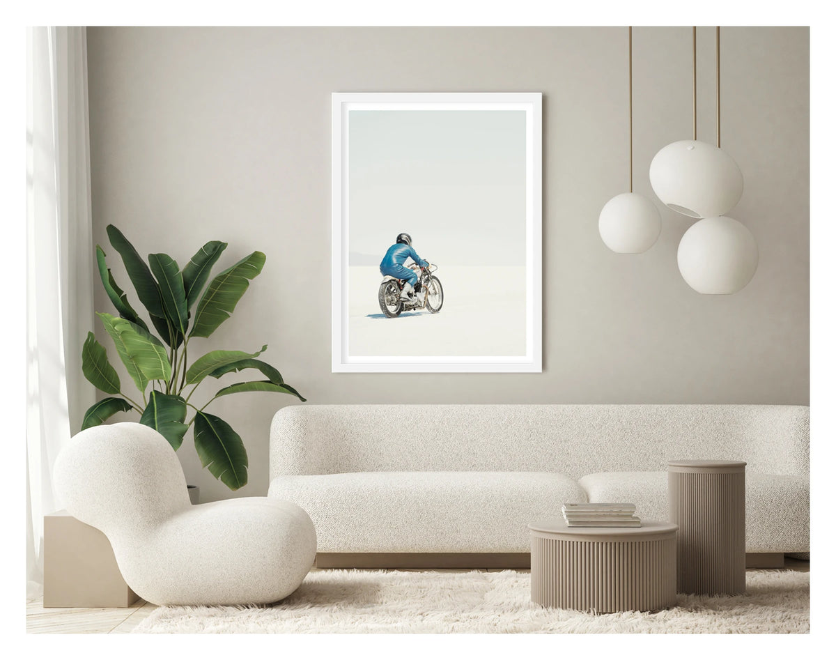 Framed in white, in a portrait orientation. A lone motorcycle racer dressed in a blue leather suit and silver helmet rides a vintage Triumph, land speed racing motorcycle across the blinding white expanse of the Bonneville salt flats, the rider and bike reduced to a small solitary figure against an almost entirely white horizon and pale sky. Shown in a contemporary living space. Fine art colour photographic print by Simon Davidson.