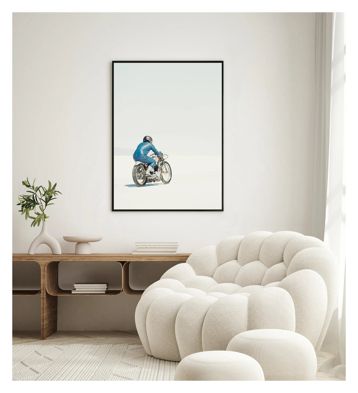 Framed in black, in a portrait orientation. A lone motorcycle racer dressed in a blue leather suit and silver helmet rides a vintage Triumph, land speed racing motorcycle across the blinding white expanse of the Bonneville salt flats, the rider and bike reduced to a small solitary figure against an almost entirely white horizon and pale sky. Shown in a contemporary living space. Fine art colour photographic print by Simon Davidson.