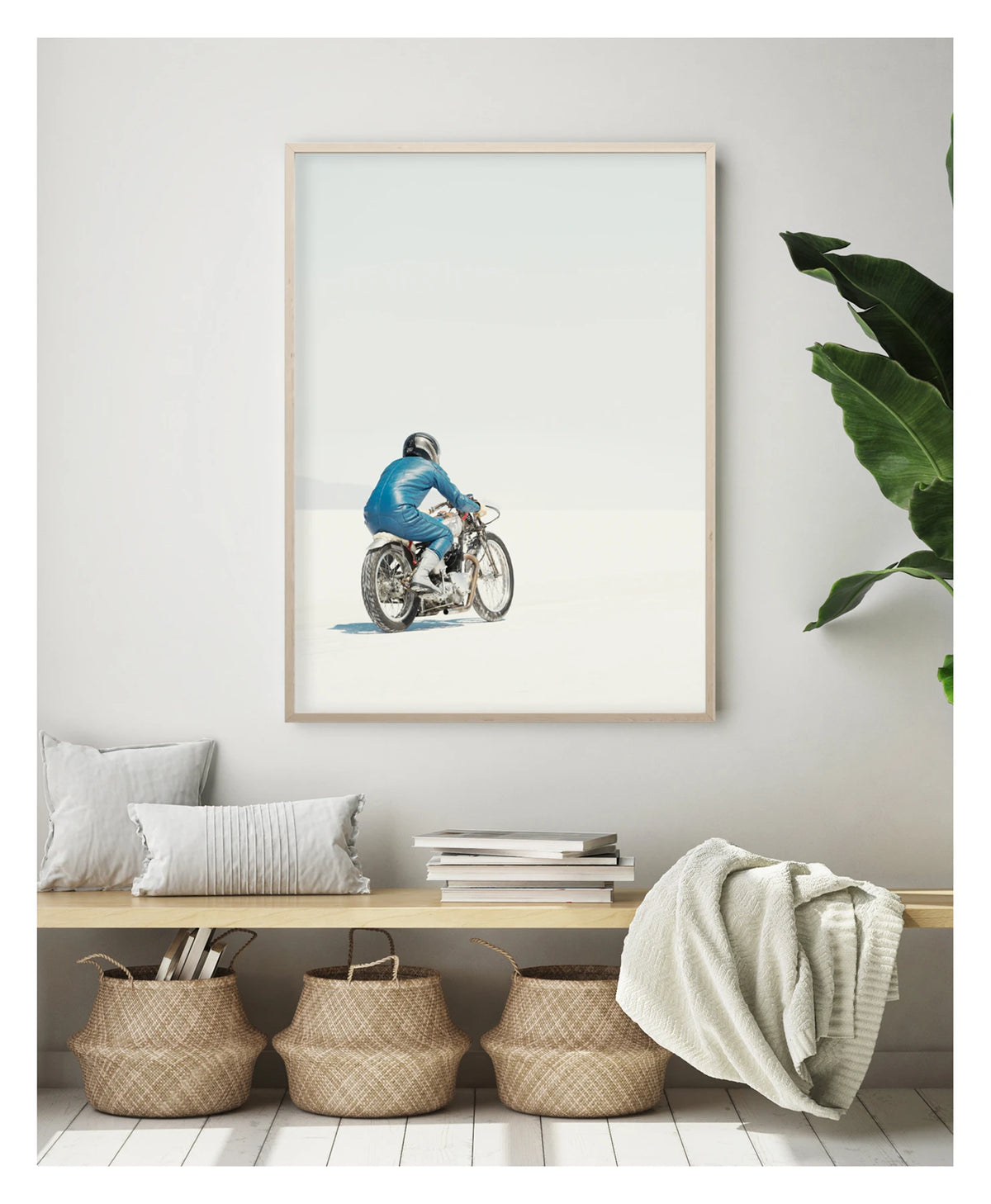 Framed in timber, in a portrait orientation. A lone motorcycle racer dressed in a blue leather suit and silver helmet rides a vintage Triumph, land speed racing motorcycle across the blinding white expanse of the Bonneville salt flats, the rider and bike reduced to a small solitary figure against an almost entirely white horizon and pale sky. Shown in a contemporary living space. Fine art colour photographic print by Simon Davidson.
