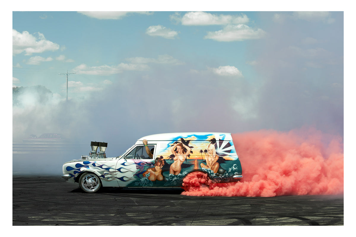 A heavily customised Australian panel van with an elaborate tropical mural and blue flame paintwork lays down a burnout under a blue sky with scattered clouds, pink tyre smoke erupting from the rear wheels and trailing behind as white smoke drifts across the left side of the frame. Supercharger protrudes through the bonnet. The lavishly painted side panel depicting a sunset beach scene in 1970s van culture glory. Fine art photographic print by Simon Davidson.