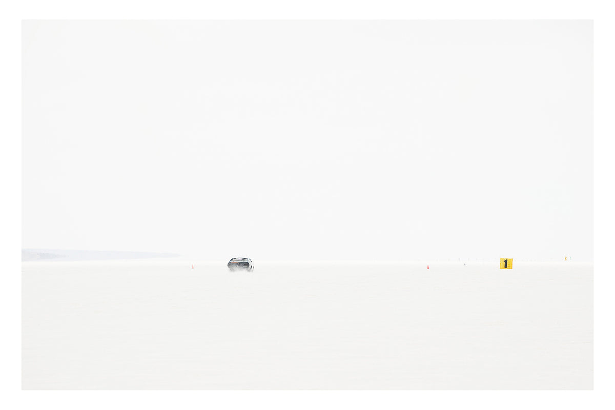 An Australian Ford XB Falcon landspeed race car appears tiny as it races down Lake Gairdner, almost entirely swallowed by the blinding white salt and pale sky. It makes a high speed run past small orange cones and a yellow number one marker. The extreme minimalism of the composition reduces the car and the entire drama of speed to a single small detail in an overwhelming expanse of white. Fine art colour photographic print by Simon Davidson.