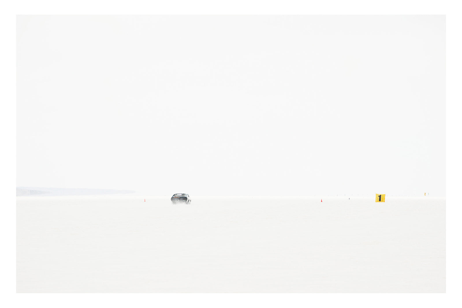 An Australian Ford XB Falcon landspeed race car appears tiny as it races down Lake Gairdner, almost entirely swallowed by the blinding white salt and pale sky. It makes a high speed run past small orange cones and a yellow number one marker. The extreme minimalism of the composition reduces the car and the entire drama of speed to a single small detail in an overwhelming expanse of white. Fine art colour photographic print by Simon Davidson.