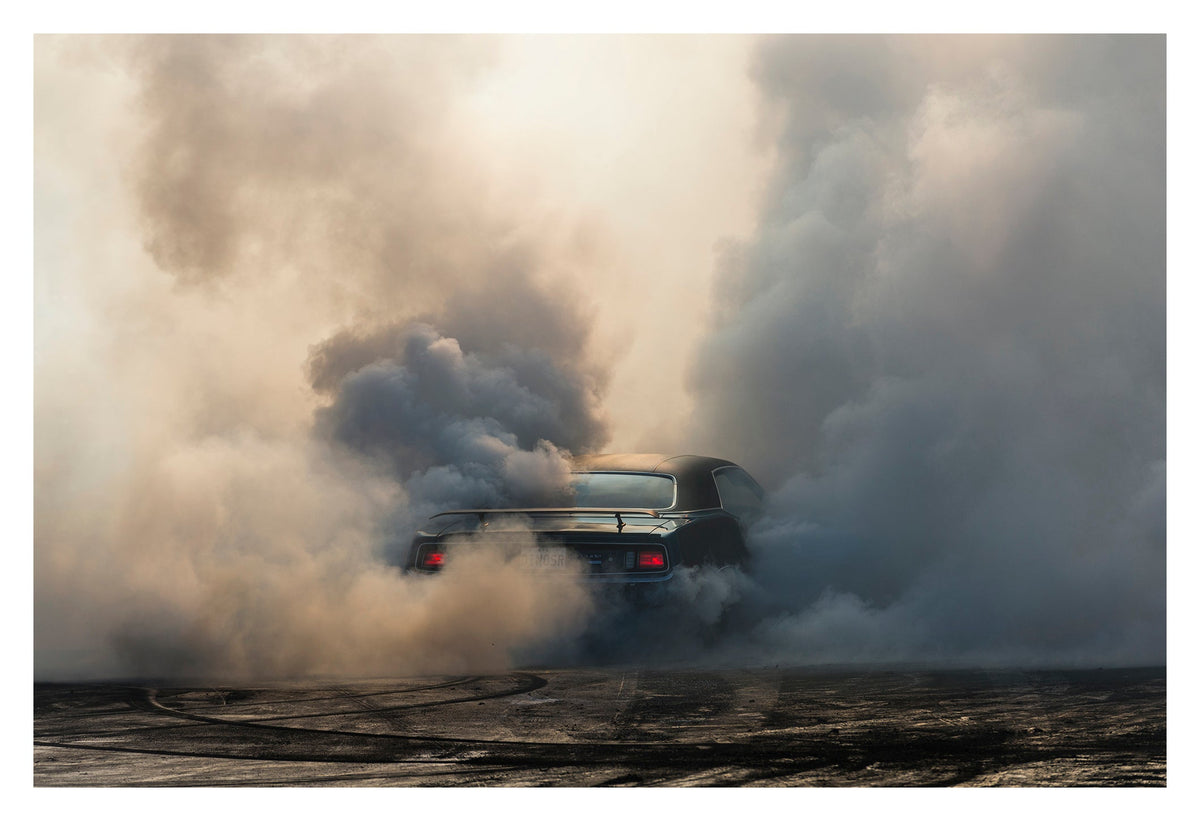 We see the rear of a black Plymouth Barracuda with a vinyl roof performing a burnout, almost entirely consumed by billowing clouds of tyre smoke that fill the frame from ground to sky. Red tail lights glow through the haze, rubber marks streak the bitumen below. Fine art colour photographic print by Simon Davidson.