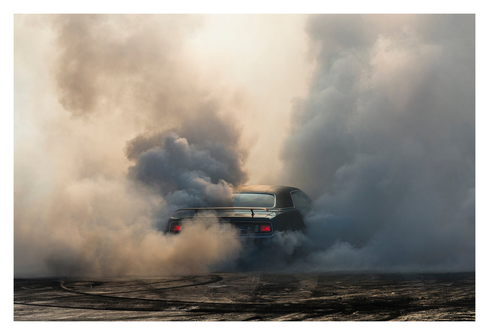 We see the rear of a black Plymouth Barracuda with a vinyl roof performing a burnout, almost entirely consumed by billowing clouds of tyre smoke that fill the frame from ground to sky. Red tail lights glow through the haze, rubber marks streak the bitumen below. Fine art colour photographic print by Simon Davidson.