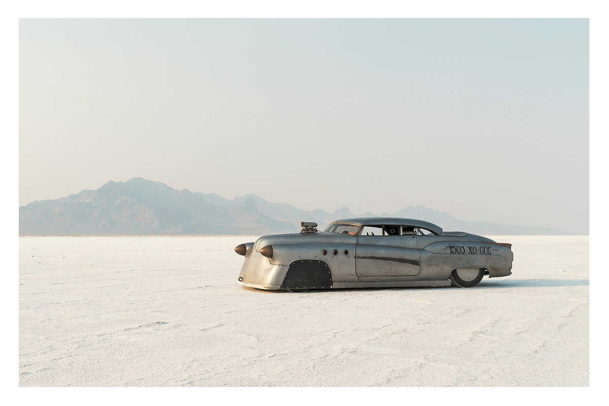 A bare metal 1950s Buick land speed racer numbered 1503 sits low on the white salt of the Bonneville Salt Flats, its body heavily modified with covered wheel arches, bullet nose fairings, porthole details and a small blower poking through the bonnet. The unpainted aluminium bodywork gleams softly against the vast white salt and hazy mountain range dissolving into a pale grey sky behind it. Fine art colour photographic print by Simon Davidson.