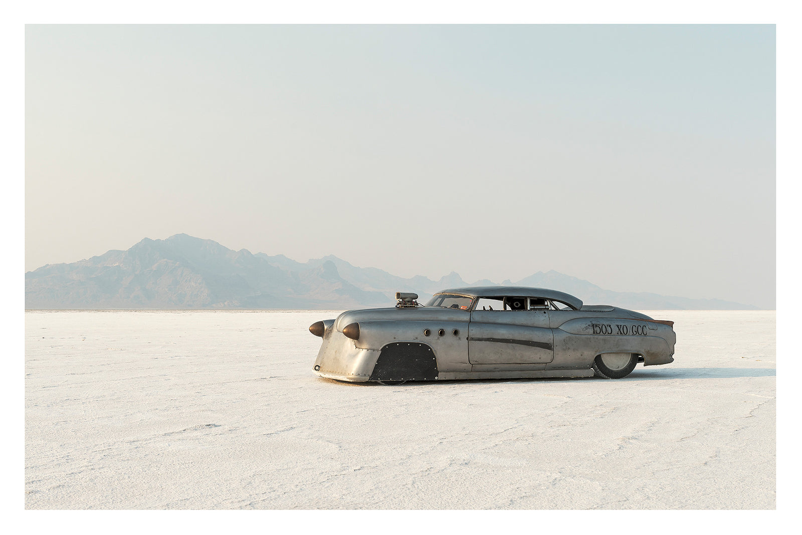 A bare metal 1950s Buick land speed racer numbered 1503 sits low on the white salt of the Bonneville Salt Flats, its body heavily modified with covered wheel arches, bullet nose fairings, porthole details and a small blower poking through the bonnet. The unpainted aluminium bodywork gleams softly against the vast white salt and hazy mountain range dissolving into a pale grey sky behind it. Fine art colour photographic print by Simon Davidson.