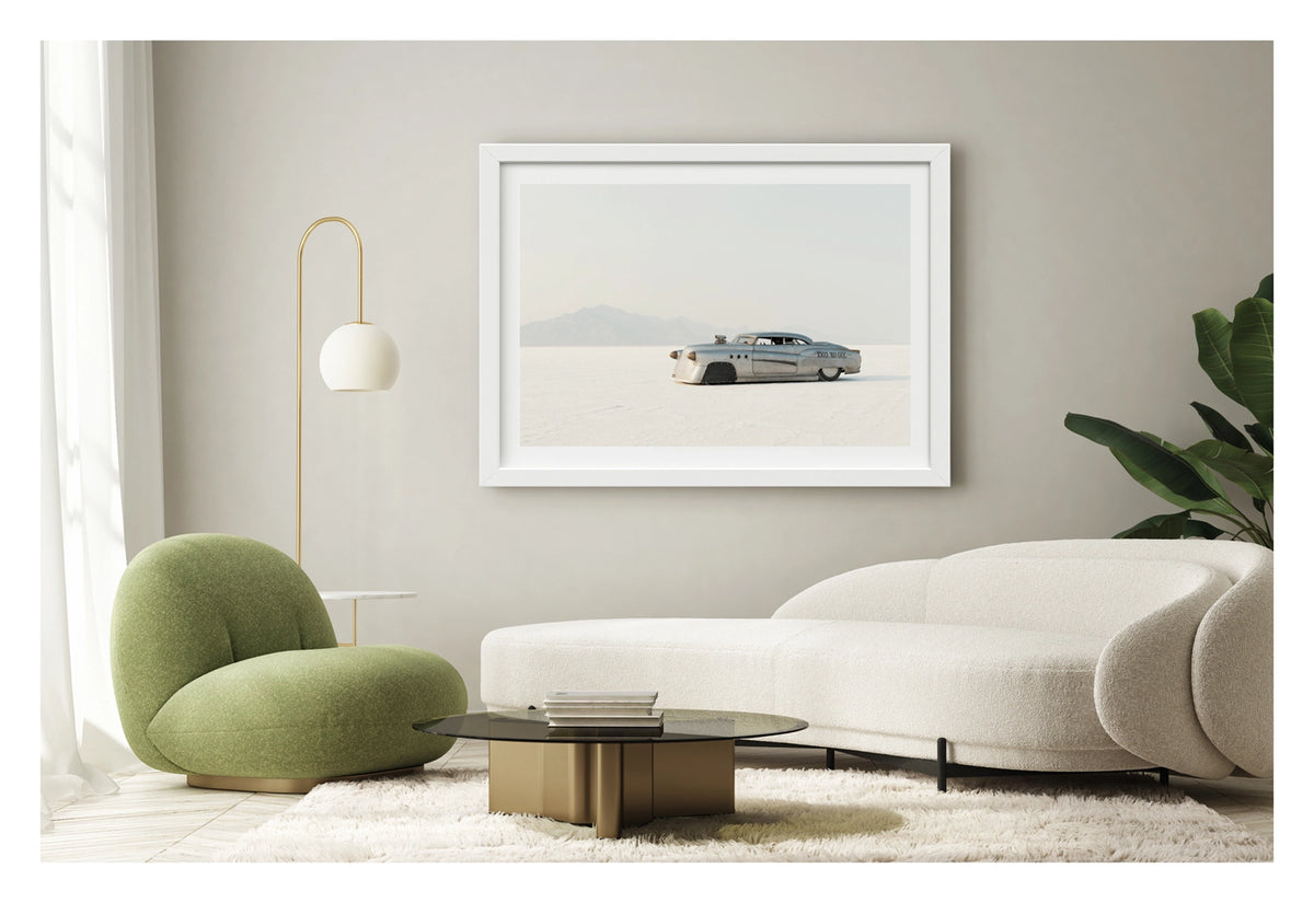 Framed in white, shown in a contemporary living space. A bare metal 1950s Buick land speed racer numbered 1503 sits low on the white salt of the Bonneville Salt Flats, its body heavily modified with covered wheel arches, bullet nose fairings, porthole details and a small blower poking through the bonnet. The unpainted aluminium bodywork gleams softly against the vast white salt and hazy mountain range dissolving into a pale grey sky behind it. Fine art colour photographic print by Simon Davidson.