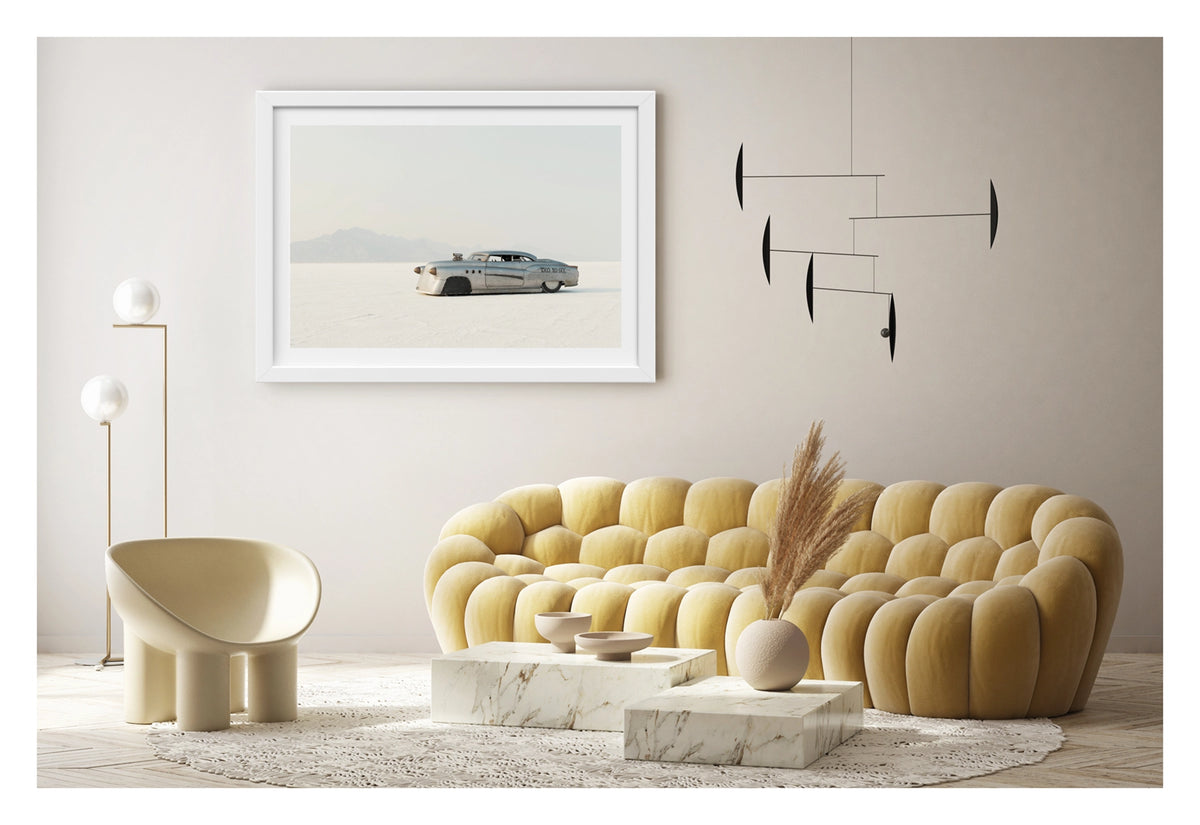 Framed in white, shown in a contemporary living space. A bare metal 1950s Buick land speed racer numbered 1503 sits low on the white salt of the Bonneville Salt Flats, its body heavily modified with covered wheel arches, bullet nose fairings, porthole details and a small blower poking through the bonnet. The unpainted aluminium bodywork gleams softly against the vast white salt and hazy mountain range dissolving into a pale grey sky behind it. Fine art colour photographic print by Simon Davidson.