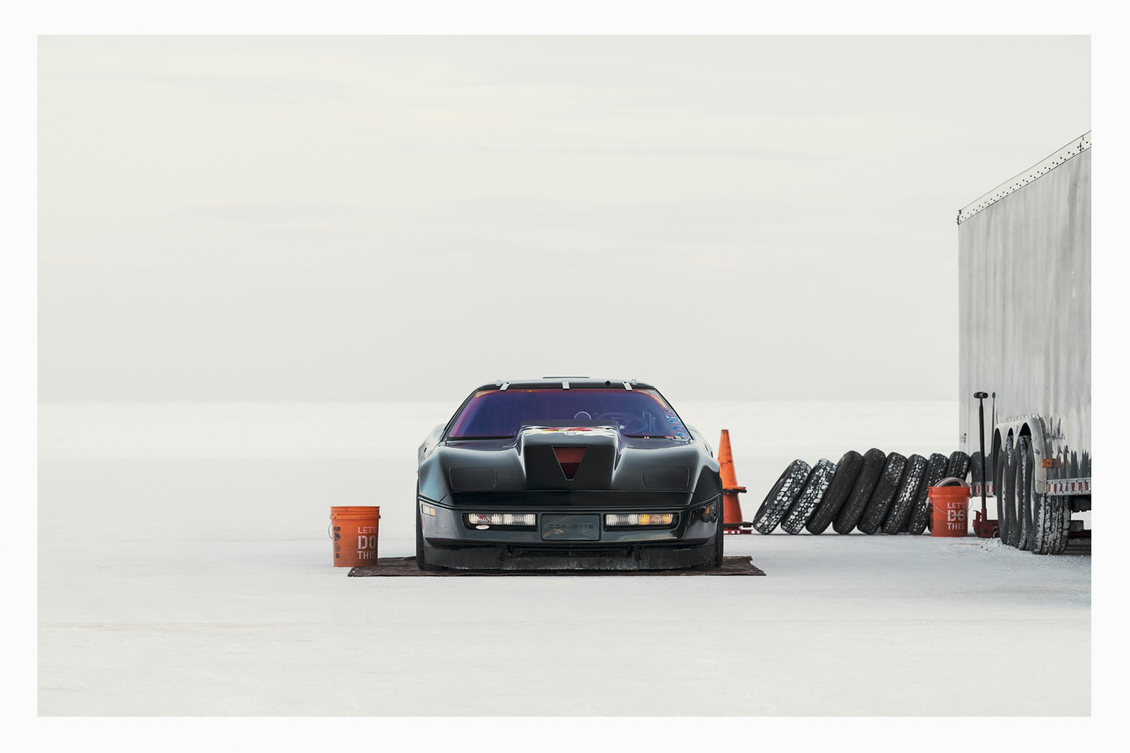 A black Chevrolet Corvette land speed racing car sits low on a ground sheet in the pits at Bonneville. Surrounded by the vast white expanse of the Salt Flats, faced directly toward the camera. Orange buckets, a row of spare tyres leaning against a trailer and an orange traffic cone are scattered around the pit area to the right, the whole scene dissolving into a featureless white horizon and sky. Fine art colour photographic print by Simon Davidson.