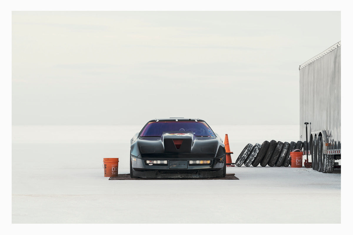 A black Chevrolet Corvette land speed racing car sits low on a ground sheet in the pits at Bonneville. Surrounded by the vast white expanse of the Salt Flats, faced directly toward the camera. Orange buckets, a row of spare tyres leaning against a trailer and an orange traffic cone are scattered around the pit area to the right, the whole scene dissolving into a featureless white horizon and sky. Fine art colour photographic print by Simon Davidson.