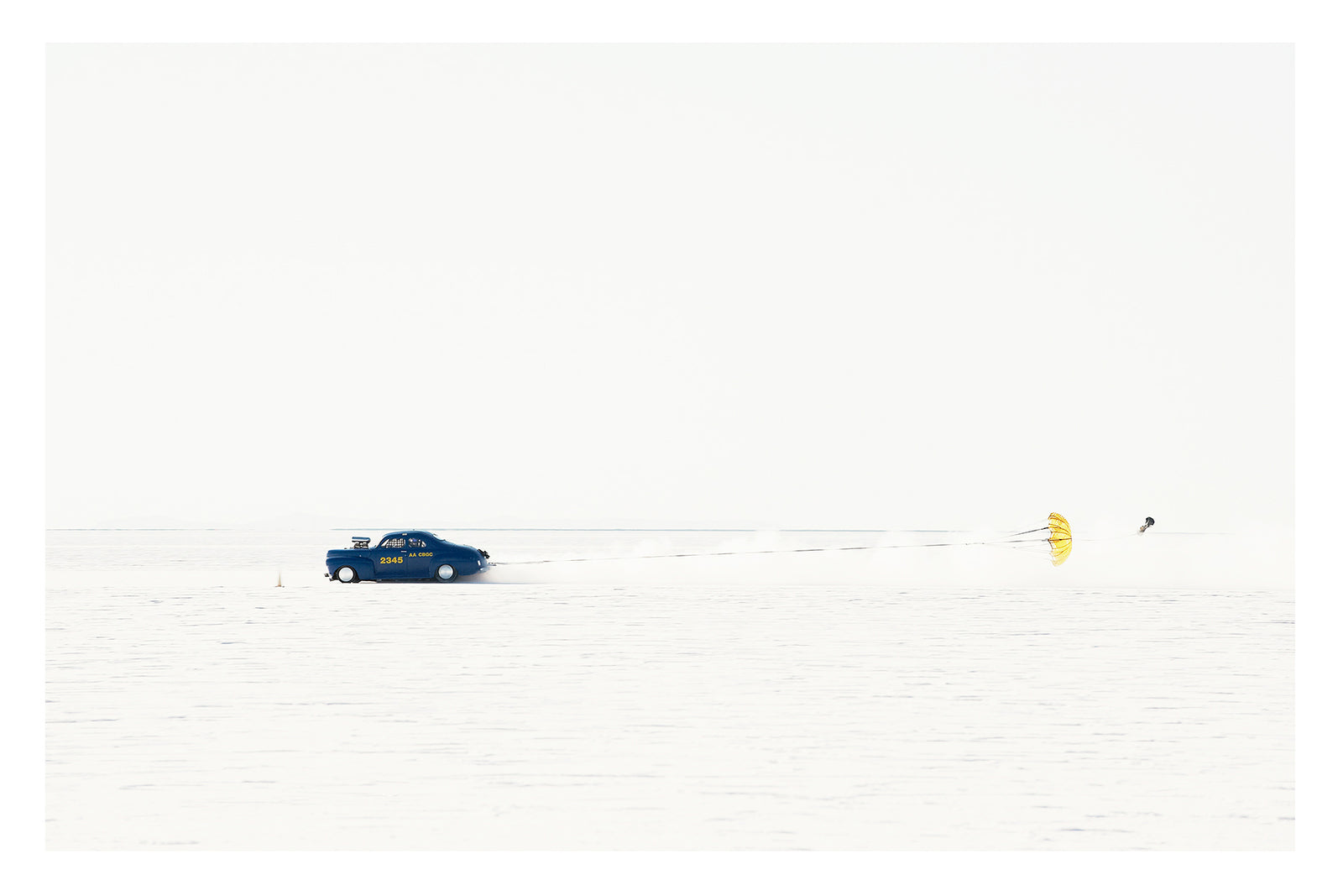 A dark blue 1940s coupe landspeed race car numbered releases a bright yellow parachute to slow down after a high speed run across the Bonneville Salt Flats, the chute billowing open behind it as a trail of disturbed salt dust follows in its wake. The car and chute sit as tiny vivid forms against the vast featureless white of the salt and sky, the scene almost entirely bleached of colour. Fine art colour photographic print by Simon Davidson.