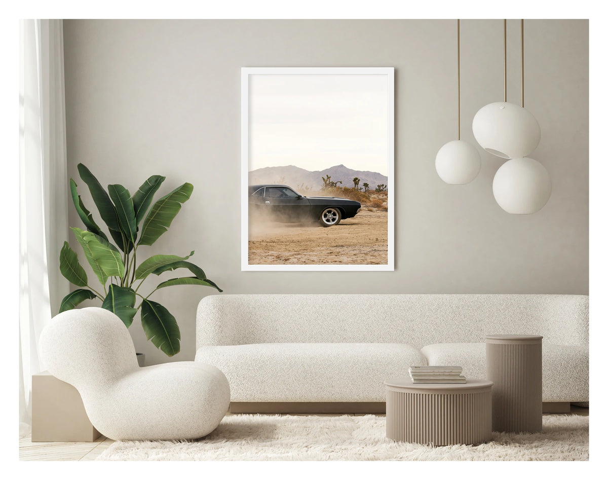 In a portrait orientation. Framed in white. A matte black first generation Dodge Challenger powers through a desert dirt surface, kicking up a low trail of dust from the rear wheels. Joshua trees and purple hue mountain ranges stretch across the horizon under a pale white sky, with the car's polished five spoke wheels and fastback roofline sharp against the hazy desert landscape. Shown in a contemporary space. Fine art colour photographic print by Simon Davidson.