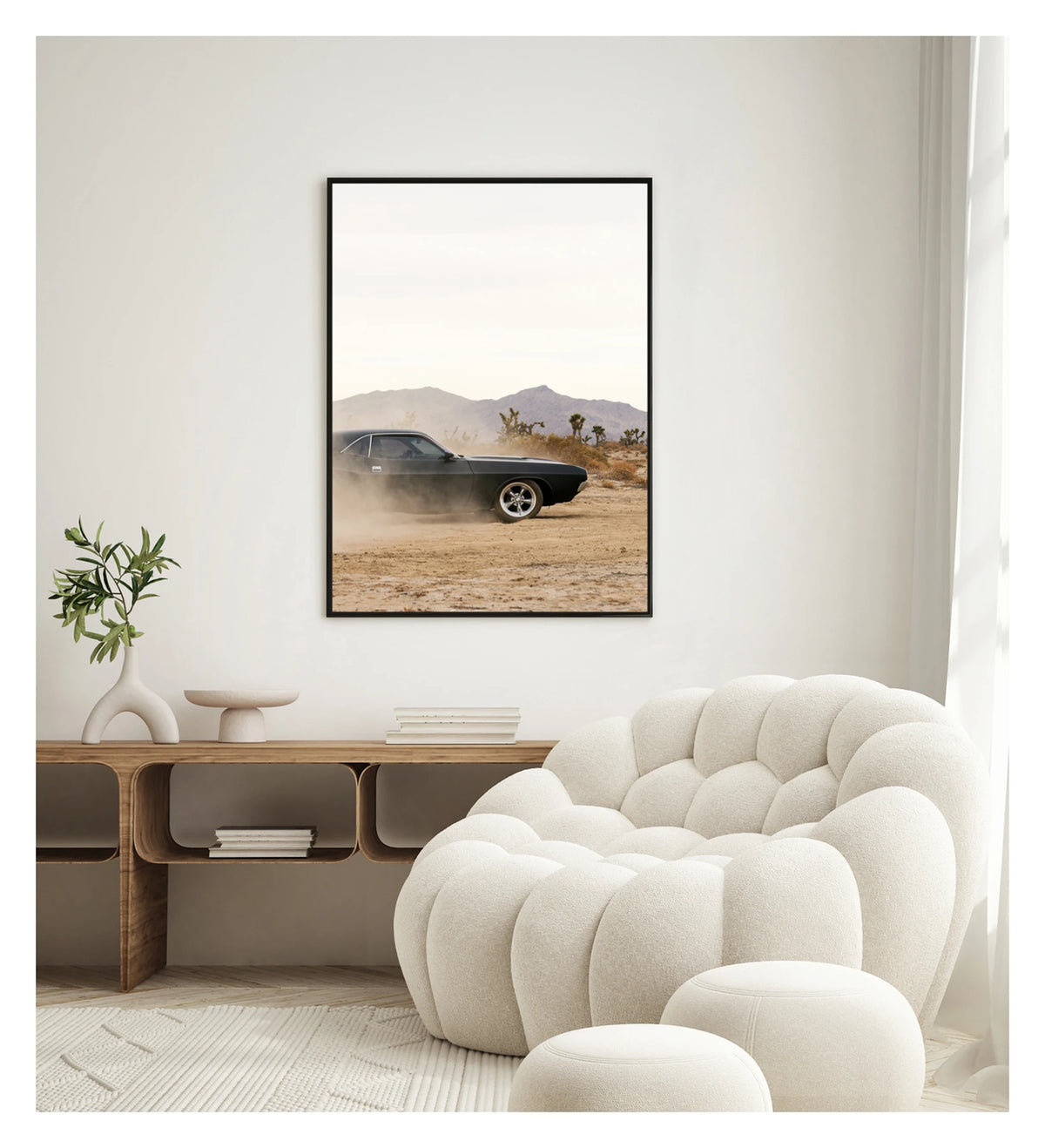 In a portrait orientation. Framed in black. A matte black first generation Dodge Challenger powers through a desert dirt surface, kicking up a low trail of dust from the rear wheels. Joshua trees and purple hue mountain ranges stretch across the horizon under a pale white sky, with the car's polished five spoke wheels and fastback roofline sharp against the hazy desert landscape. Shown in a contemporary space. Fine art colour photographic print by Simon Davidson.