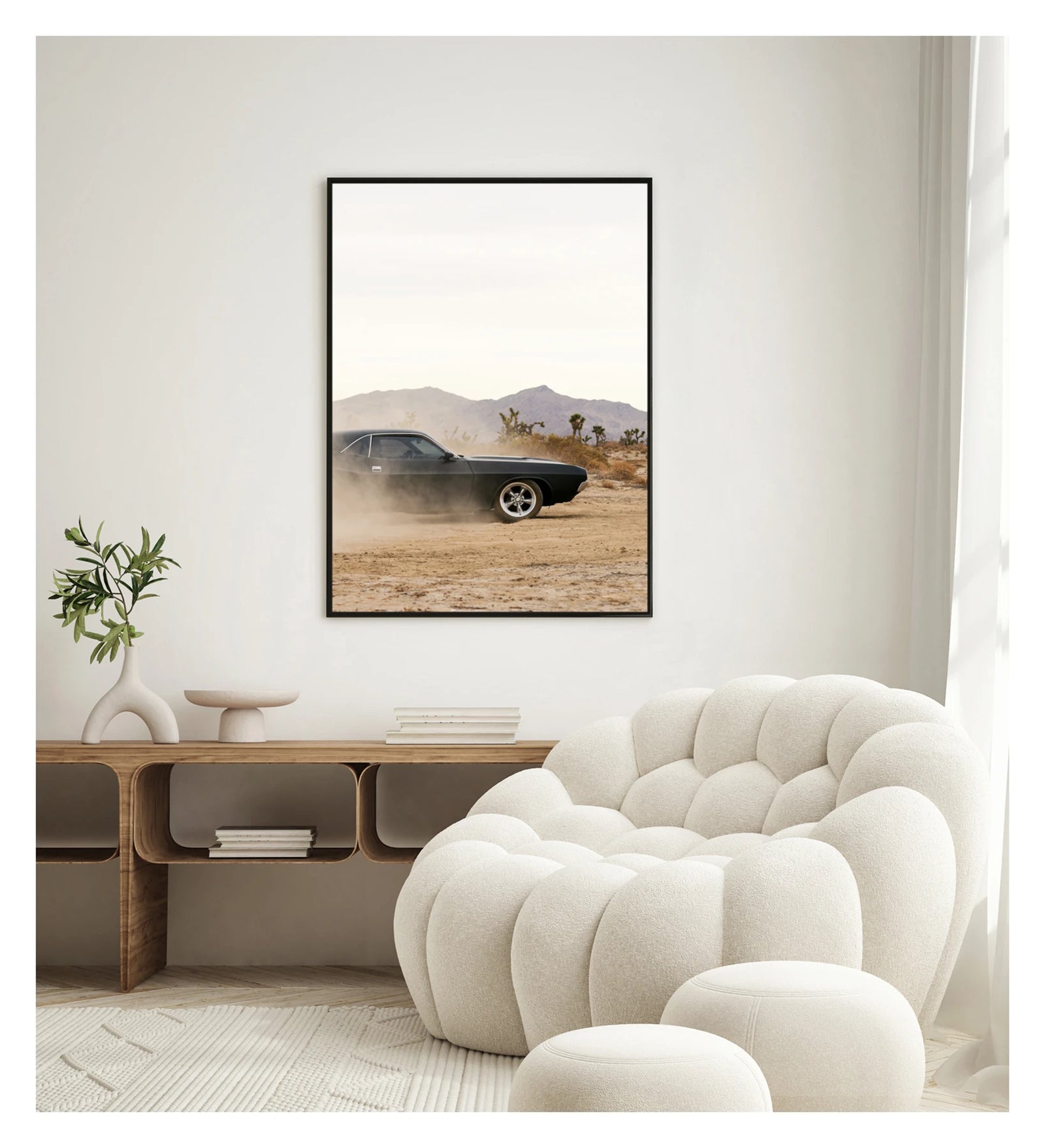 In a portrait orientation. Framed in black. A matte black first generation Dodge Challenger powers through a desert dirt surface, kicking up a low trail of dust from the rear wheels. Joshua trees and purple hue mountain ranges stretch across the horizon under a pale white sky, with the car's polished five spoke wheels and fastback roofline sharp against the hazy desert landscape. Shown in a contemporary space. Fine art colour photographic print by Simon Davidson.