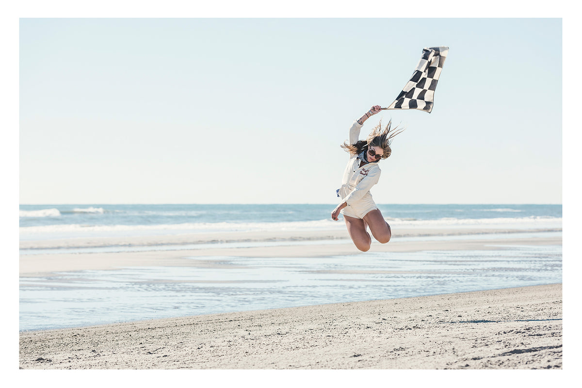 A young woman leaps high into the air on a wide empty beach, waving a black and white chequered racing flag above her head as her hair flies in the wind. Wearing a white shirt and shorts, sunglasses and a neck scarf, she is captured mid jump with knees bent against a pale blue sky and gentle surf rolling in behind her. Fine art colour photographic print by Simon Davidson.