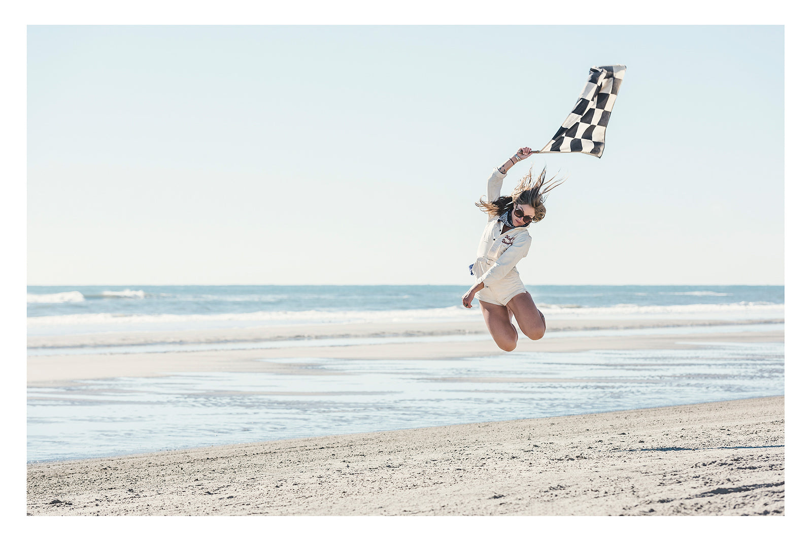 A young woman leaps high into the air on a wide empty beach, waving a black and white chequered racing flag above her head as her hair flies in the wind. Wearing a white shirt and shorts, sunglasses and a neck scarf, she is captured mid jump with knees bent against a pale blue sky and gentle surf rolling in behind her. Fine art colour photographic print by Simon Davidson.